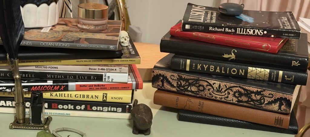 Two stacks of books on her bedside table: Malcolm X, Kahlil Gibran, Leonard Cohen, Joseph Campbell, Ocean Vuong, The Kybalion, The Upanishads, Illusions, The Art of War.
