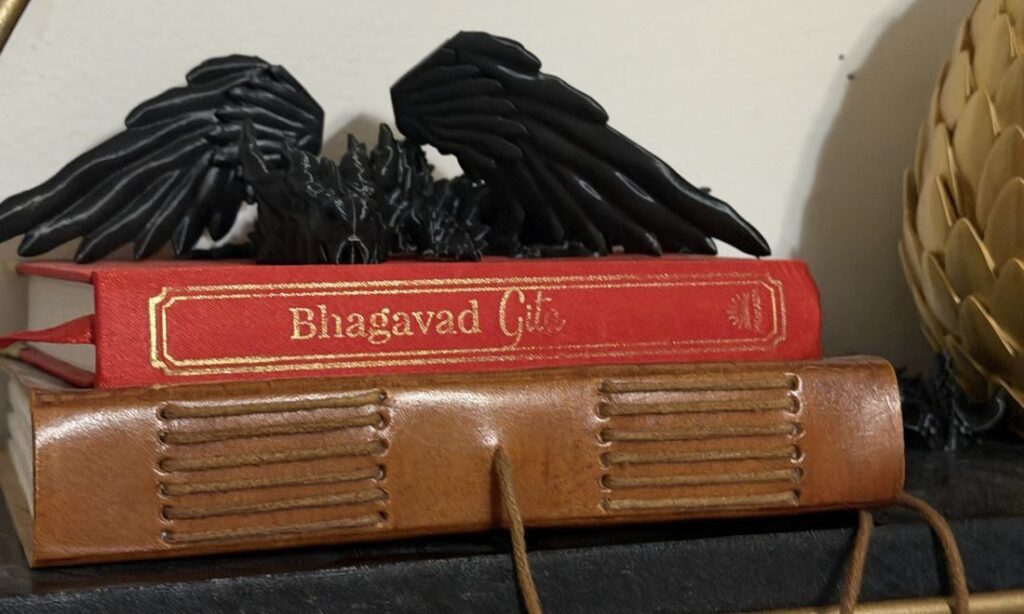 A black articulated dragon spreading its wings on top of a red Bhagavad Gita and a leather-bound journal.