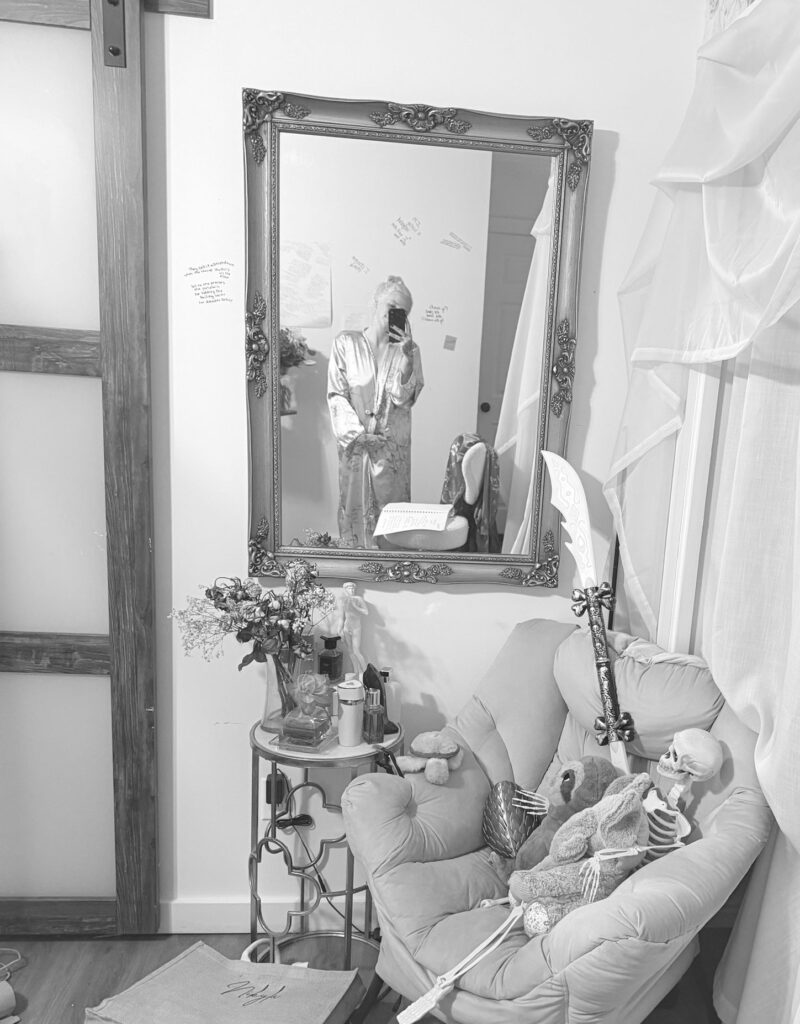 Black-and-white photograph of a gold mirror reflecting Nikyla, her phone held over her face, the pink chair beside her.