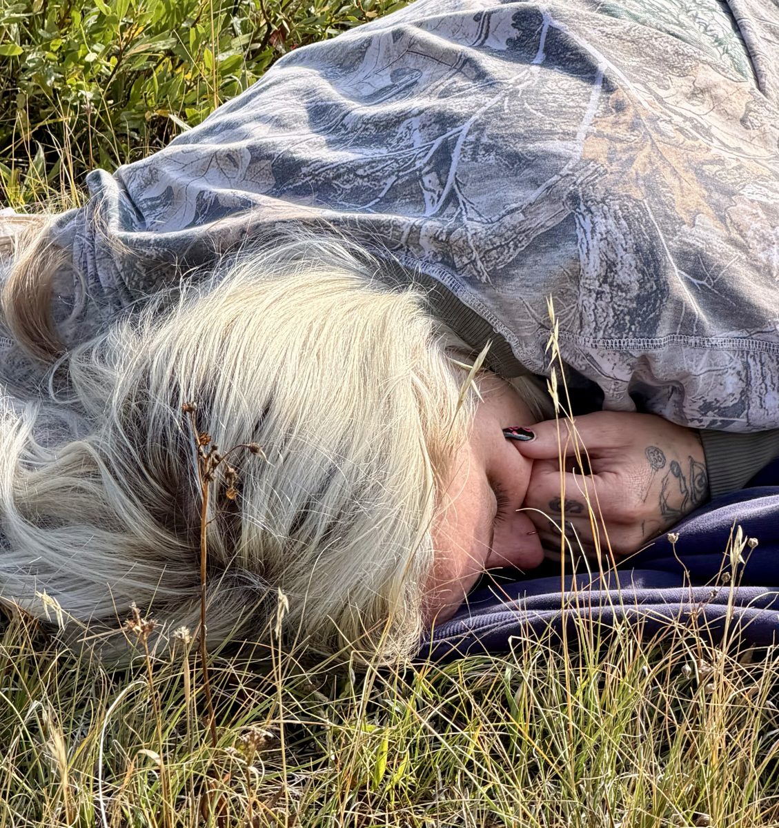 Close-up of Nikyla asleep in the grass, face tucked into her tattooed hand, hood pulled over her blonde hair.
