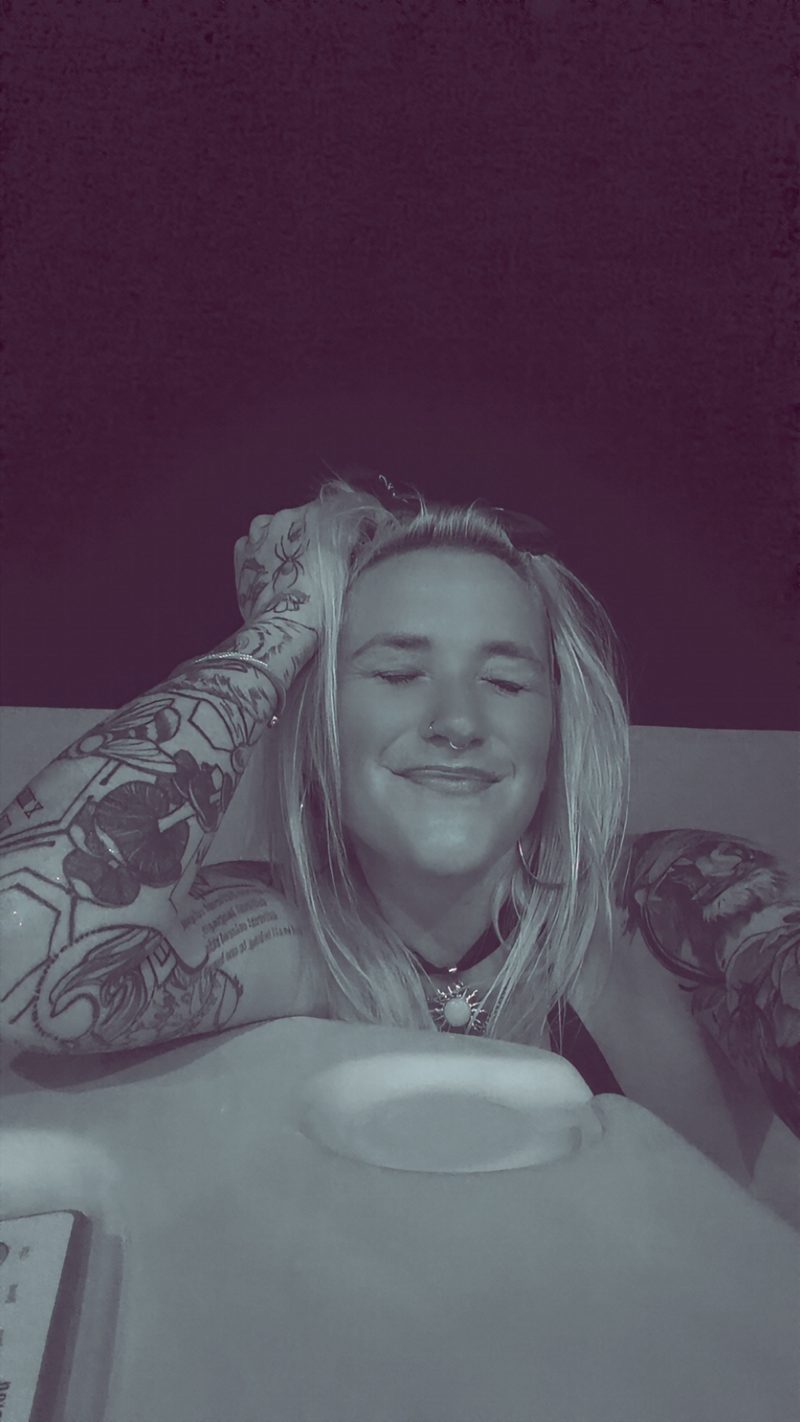 Nikyla smiling with her eyes closed, hand in her hair, tattooed arm resting on a white tub, in purple-tinted light.