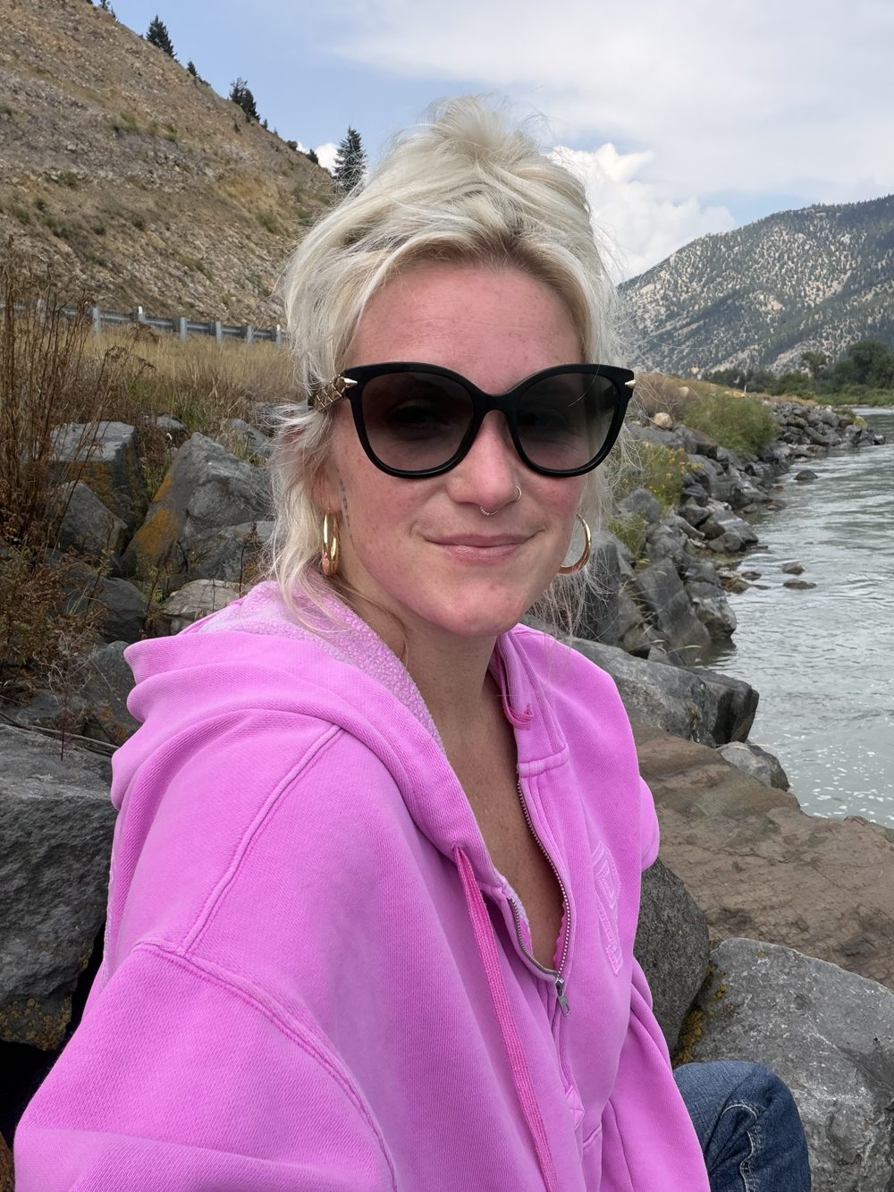 Nikyla in a pink hoodie and sunglasses, smiling, sitting on the rocks by a mountain river.