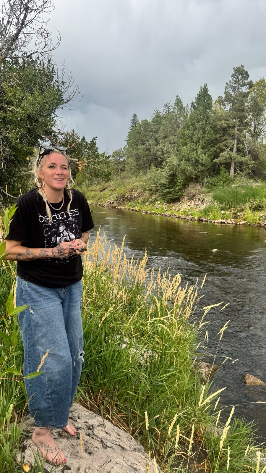 Nikyla barefoot on a rock by a river, braids and sunglasses, winking at the camera.